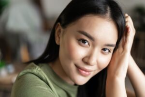 Close up portrait of tender asian girl with glowing natural skin, smiling sensual at camera, sitting
