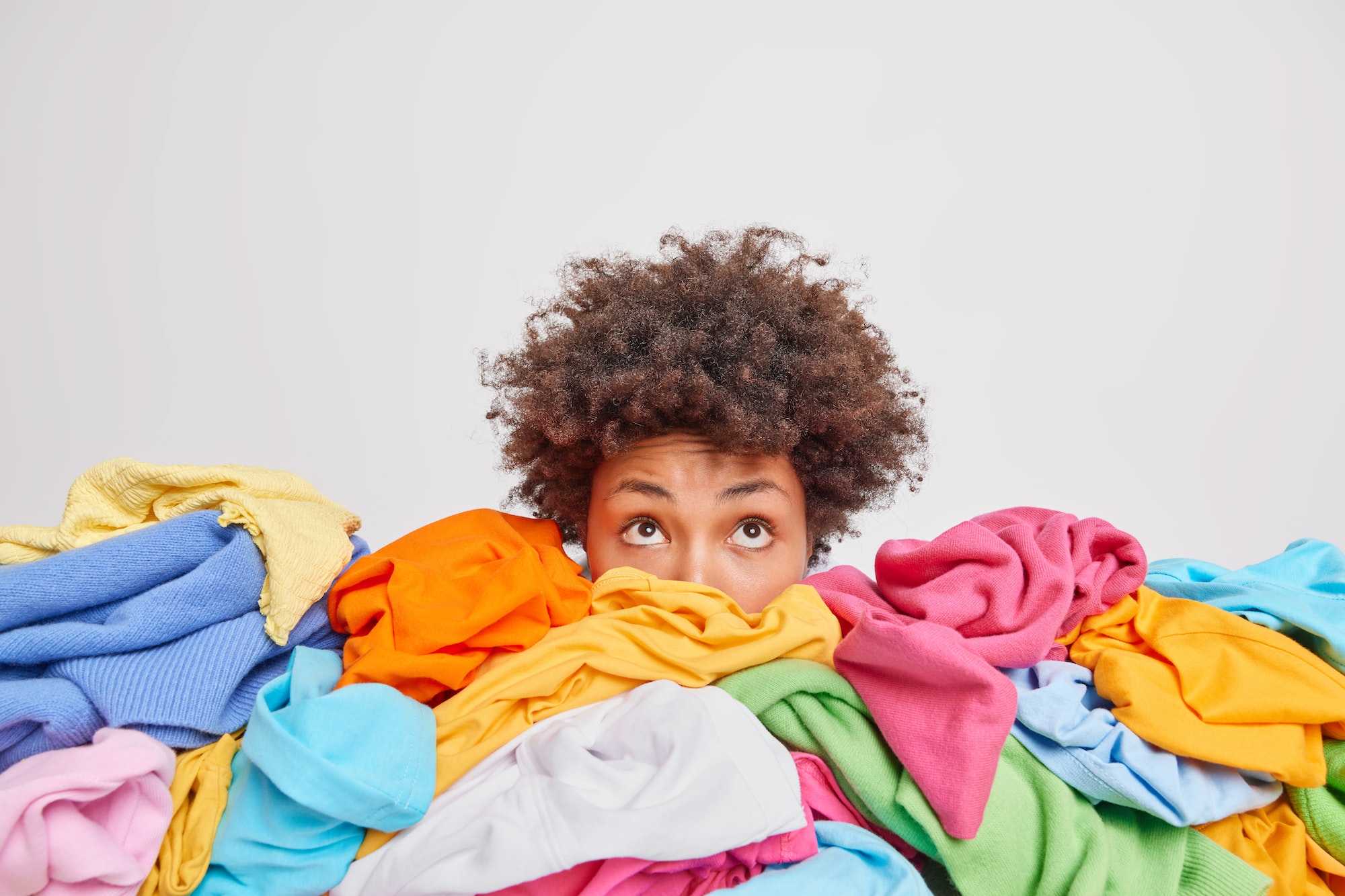 Wondered curly haired ethnic woman focused above surrounded by multicolored laundry cluttered with c