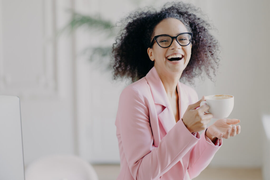 Overjoyed woman laughs happily while drinks hot coffee or latte, wears transparent spectacles