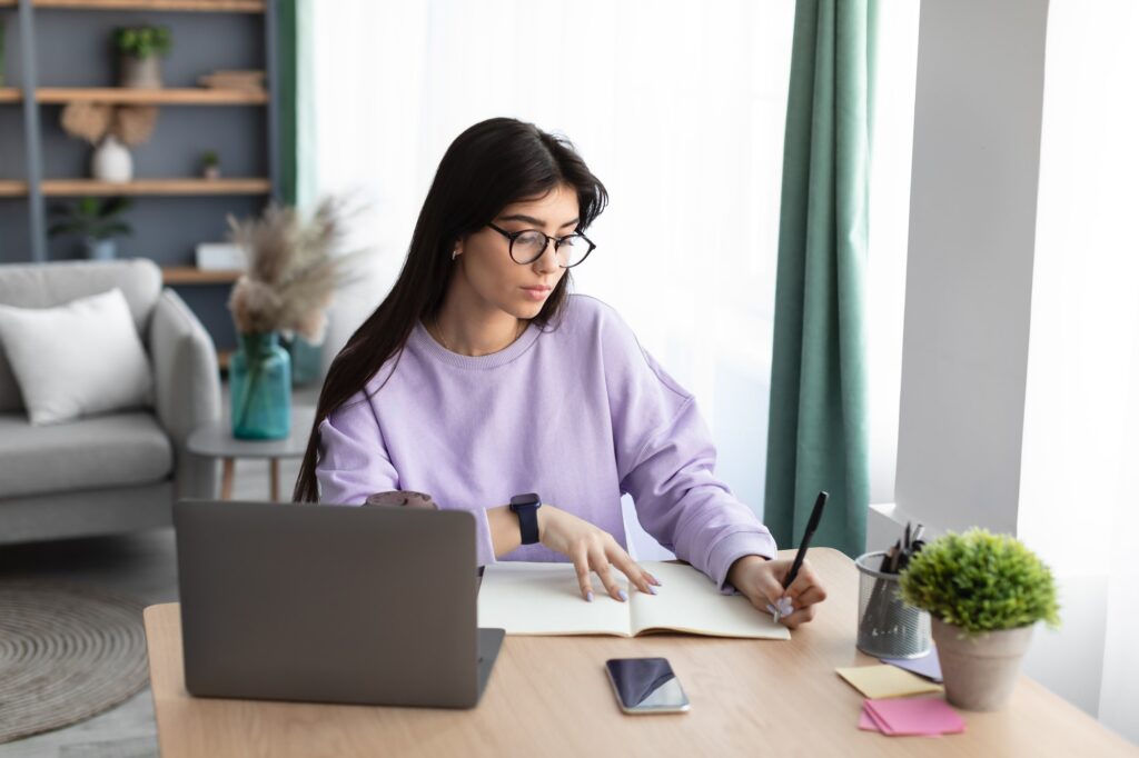 Woman sitting at desk, using computer and writing in notebook Balancing Work and Wellness