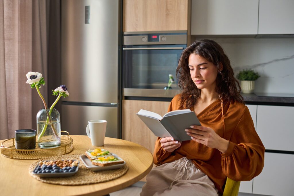 Young relaxed woman in casualwear reading book while sitting by kitchen table Balancing Work and Wellness