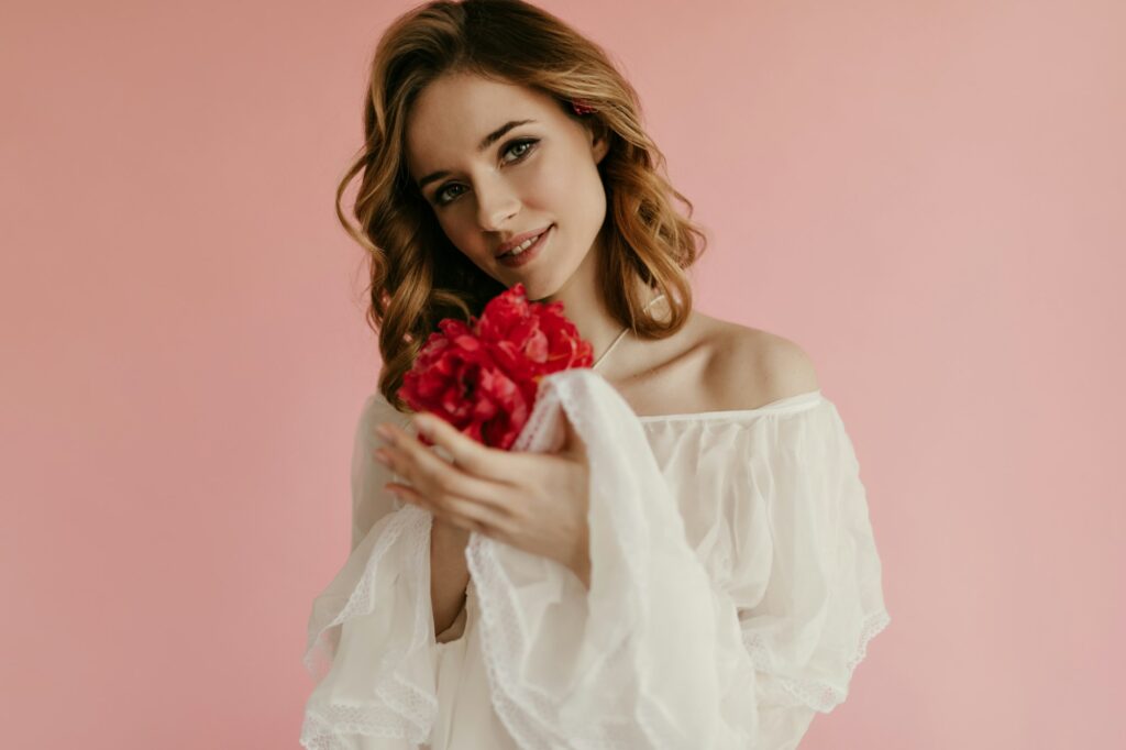 Cute curly girl with delicate wedding makeup enjoys smell of bouquets of red tulips on pink background. 