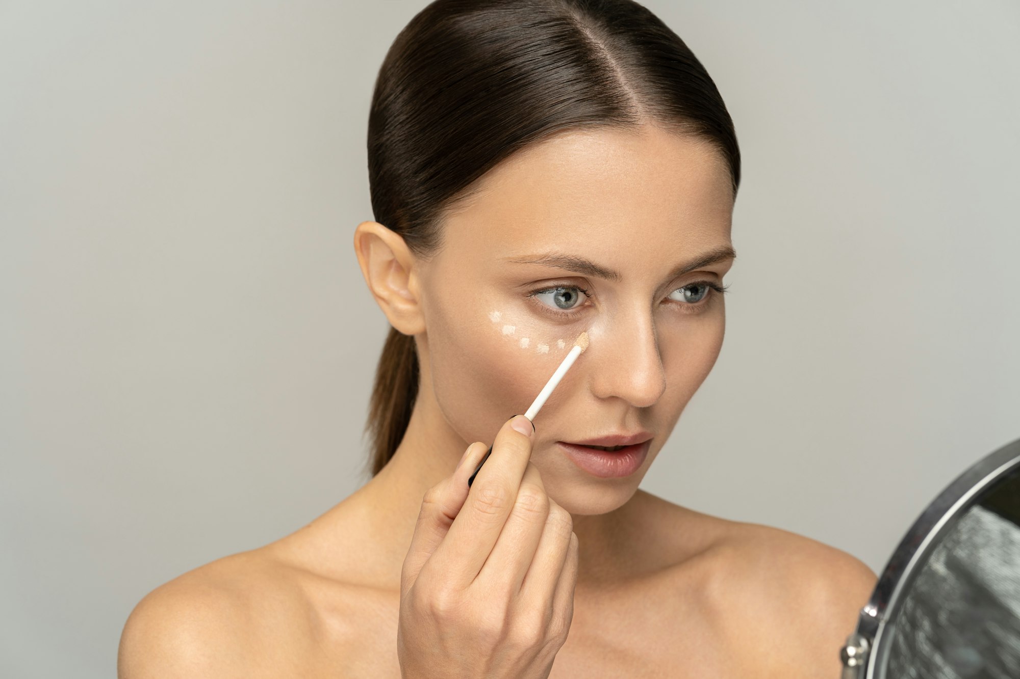Woman with natural makeup applying concealer on flawless fresh skin, doing make up looking at mirror