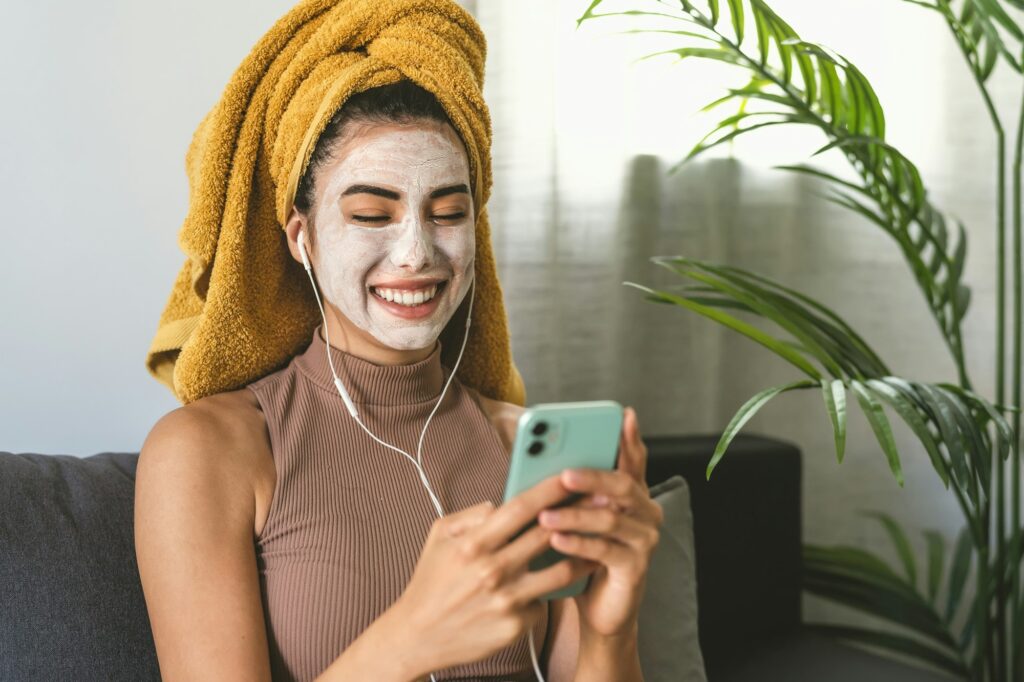 Young woman using mobile smartphone while having skin care spa day at home DIY Facial at Home