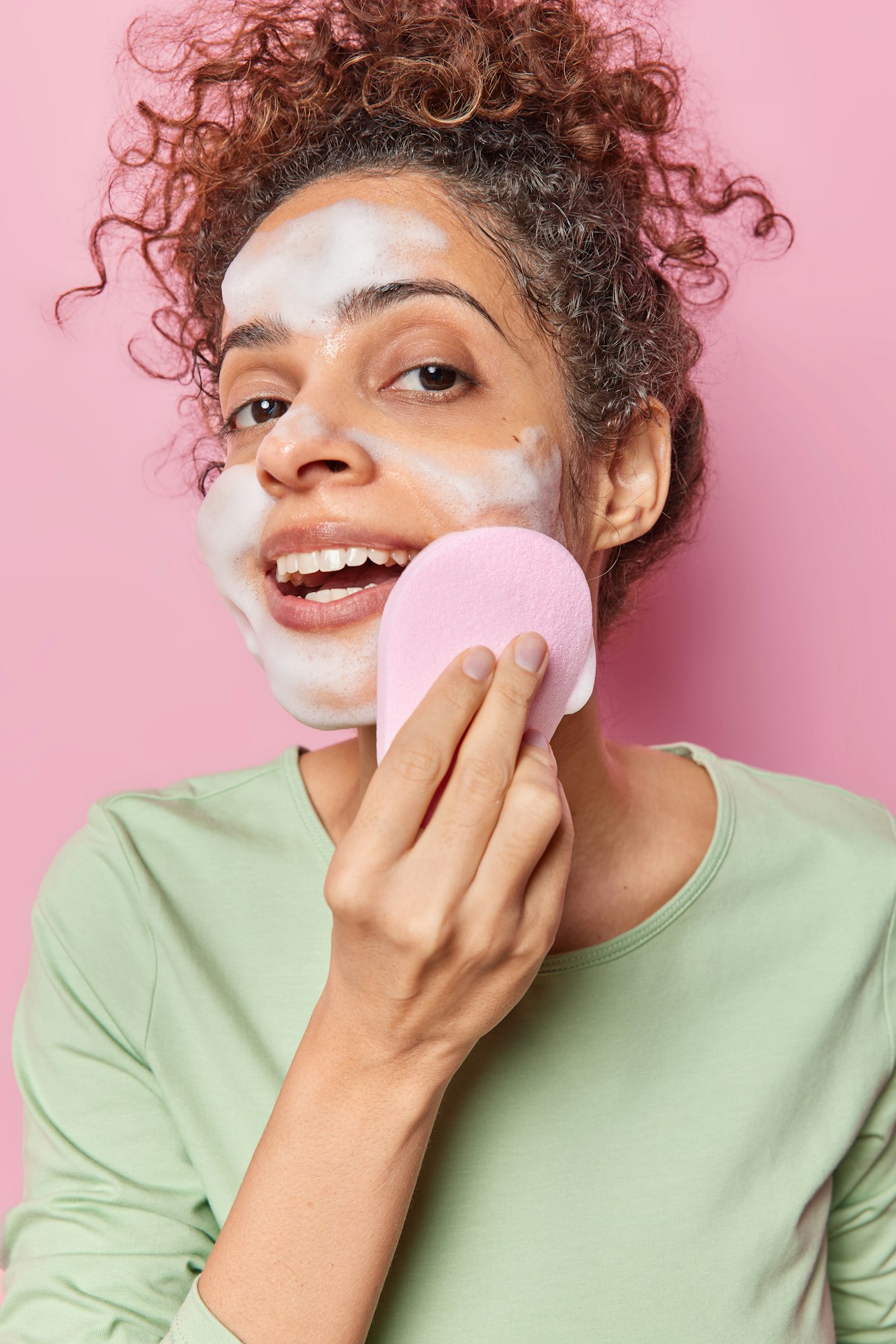 Vertical shot of curly haired woman washes face with sponge applies soap bubble wants to cleanse ans